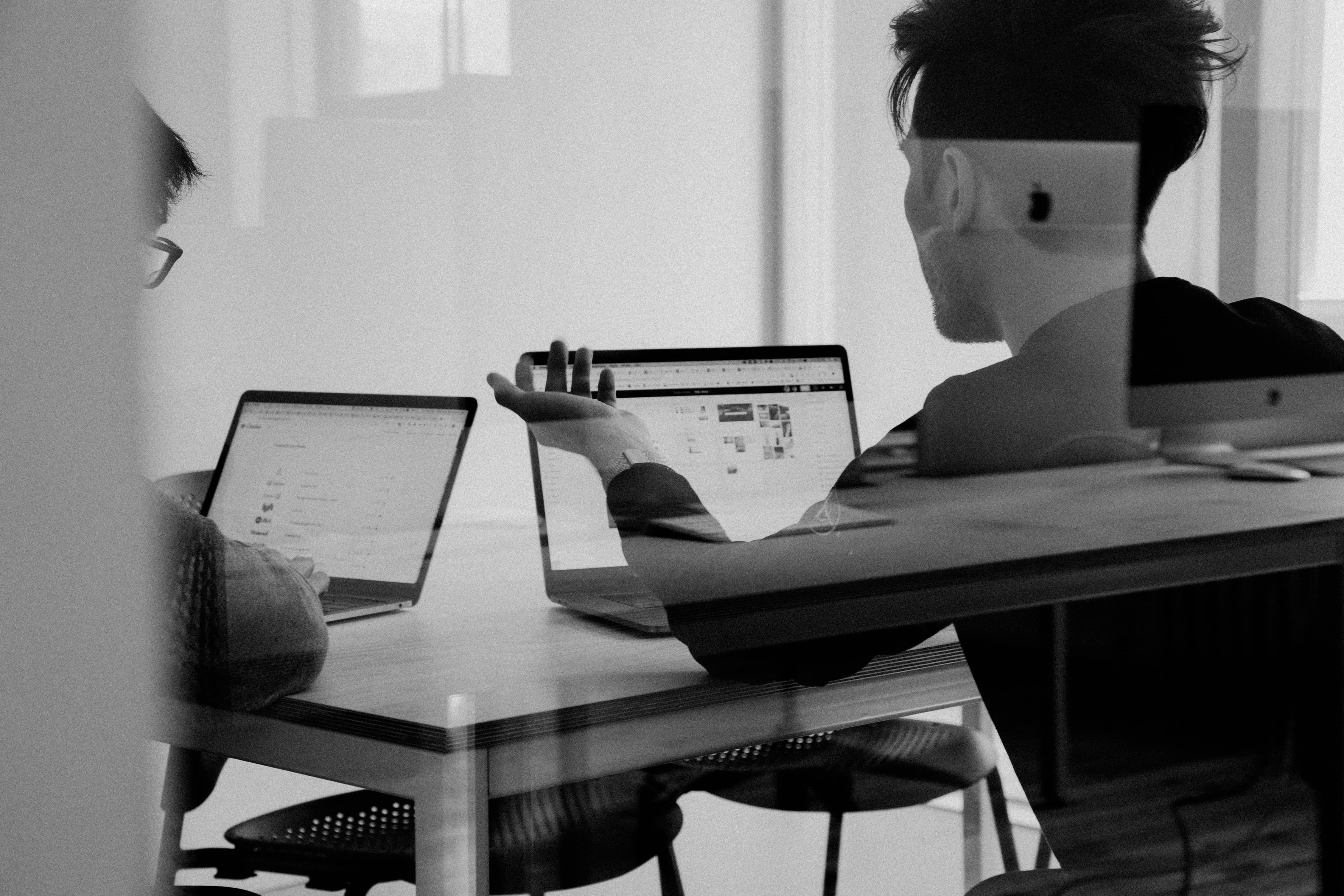Two people working on laptops at a desk, viewed through a reflective glass partition.